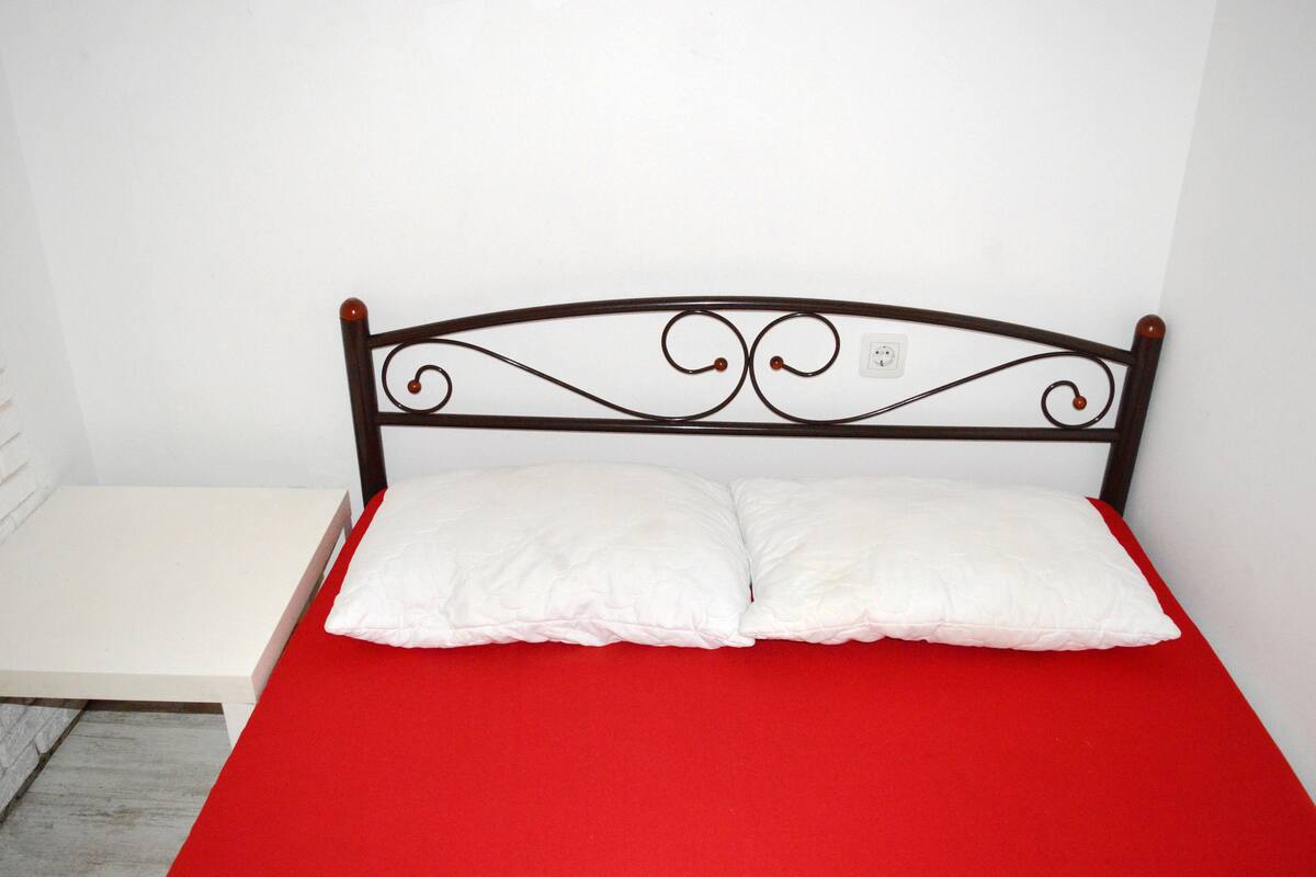 A bed with a red mattress and white pillows, a white nightstand, and a white wall.