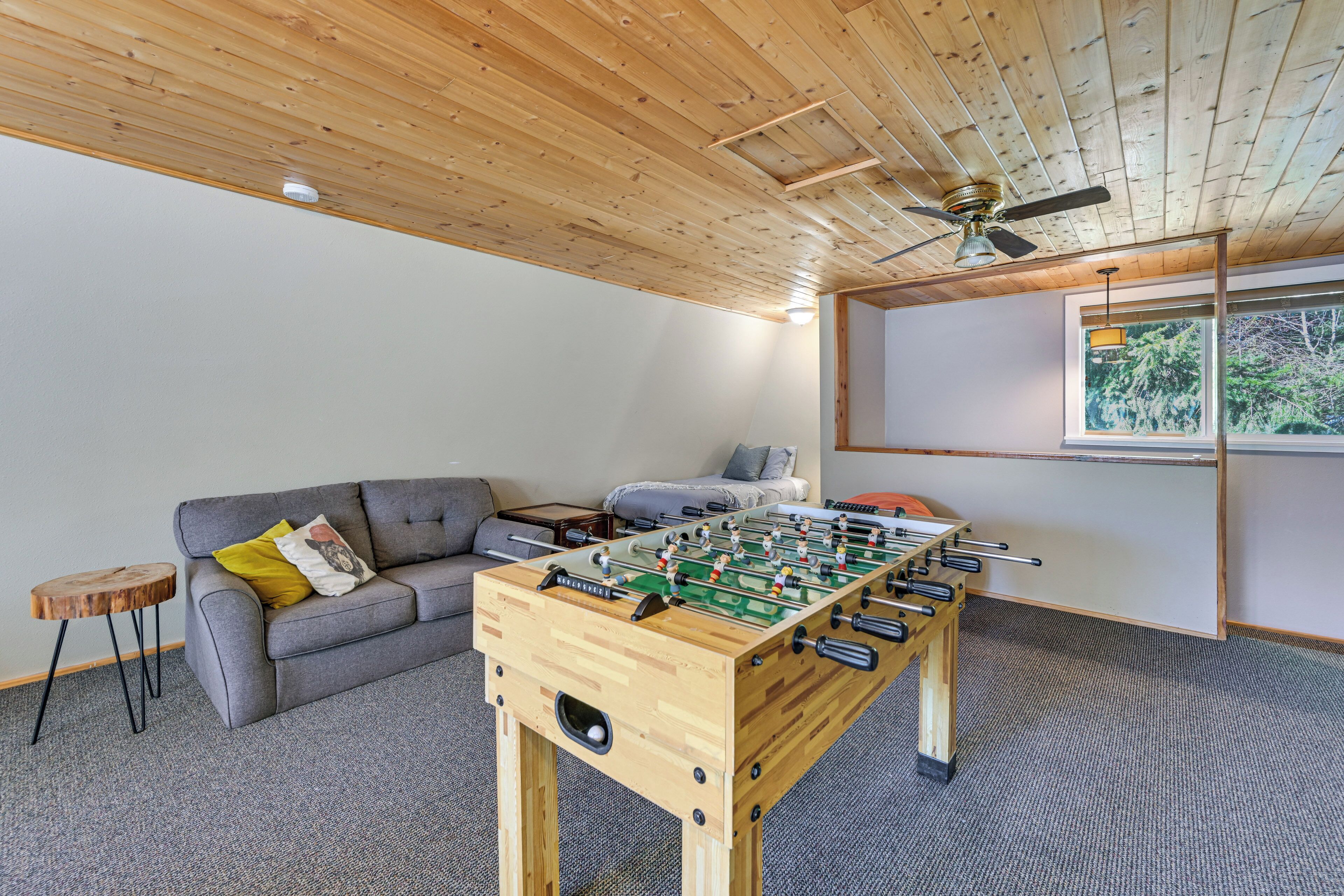 Una habitación con mesa de fútbol de mesa, un sofá gris, techo de madera y ventana con vistas.
