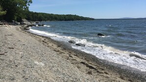 A pebble beach with a rocky shoreline, a staircase leading to a wooden structure, and a forested hill in the background. - Ocean-Beach Front Cottage on Blue Hill Bay w/ Mt Desert Island on the Horizon... (Surry)