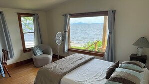 A bedroom with a bed, a chair, a fan, a lamp, and a window with a view of the sea. - Ocean-Beach Front Cottage on Blue Hill Bay w/ Mt Desert Island on the Horizon... (Surry)