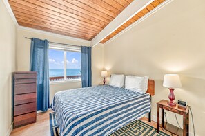 A hotel room with a bed, a dresser, a nightstand, a lamp, and a window with a view of the sea.
