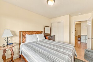 A bedroom with a wooden headboard, a bed with a striped bedspread, a bedside table with a lamp, a dresser, and a mirror.