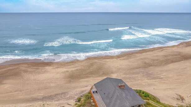 Une maison de plage avec un toit en pente, située sur une plage de sable, avec l’océan et ses vagues en arrière-plan.