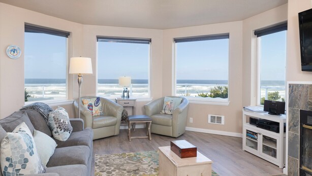 A living room with a sofa, two armchairs, a coffee table, and a fireplace. There are large windows with a view of the ocean.