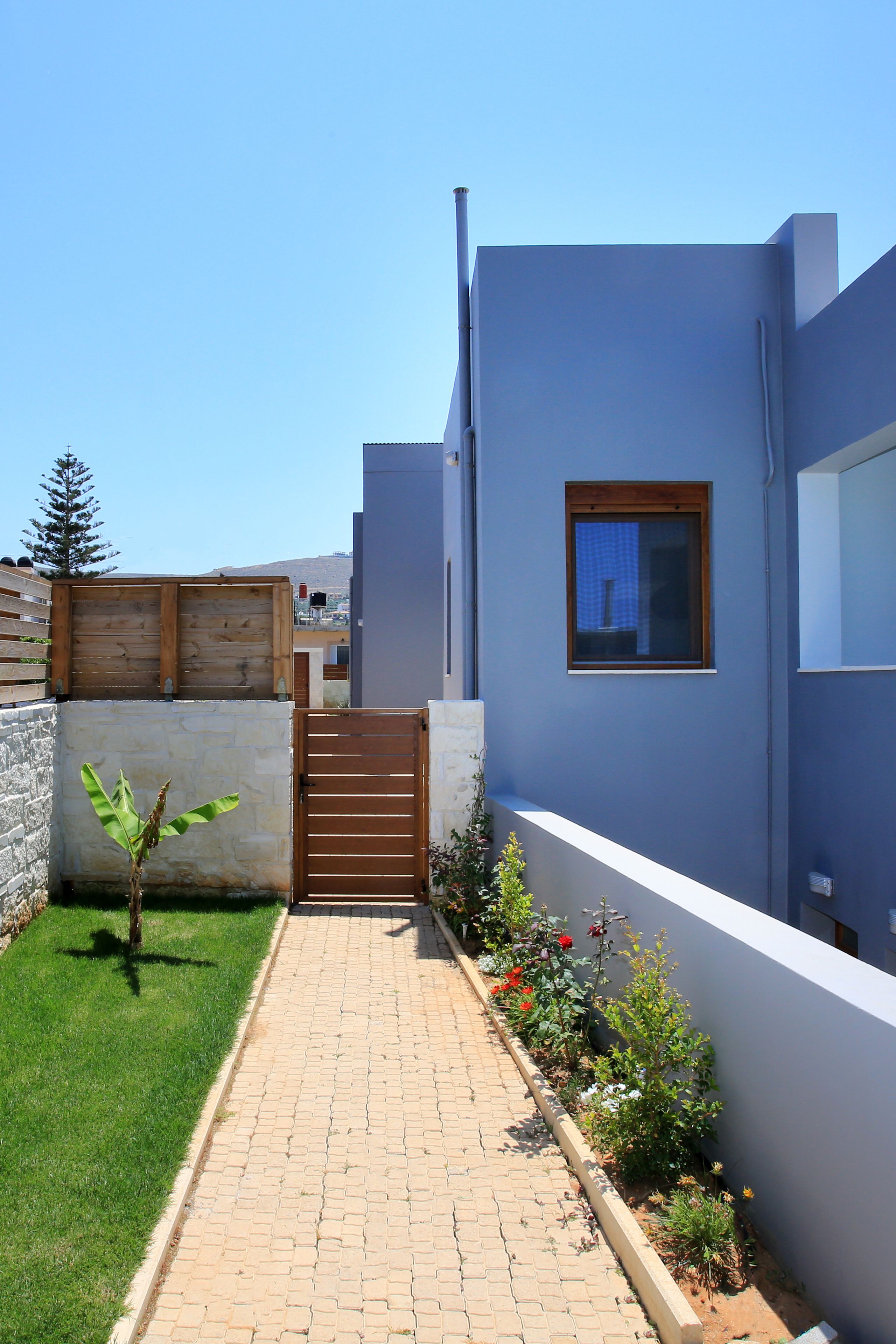 A modern building with a paved pathway, a wooden gate, and a small garden with various plants and flowers.