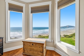 Quarto com três grandes janelas com vista para uma praia, um baú de madeira e uma parede branca.