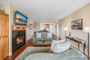 A living room with a fireplace, a television mounted on a wooden mantel, and a sofa with patterned cushions. - Panoramic Pacific Views at this 2nd floor luxury oceanfront condo, beach access (Newport)