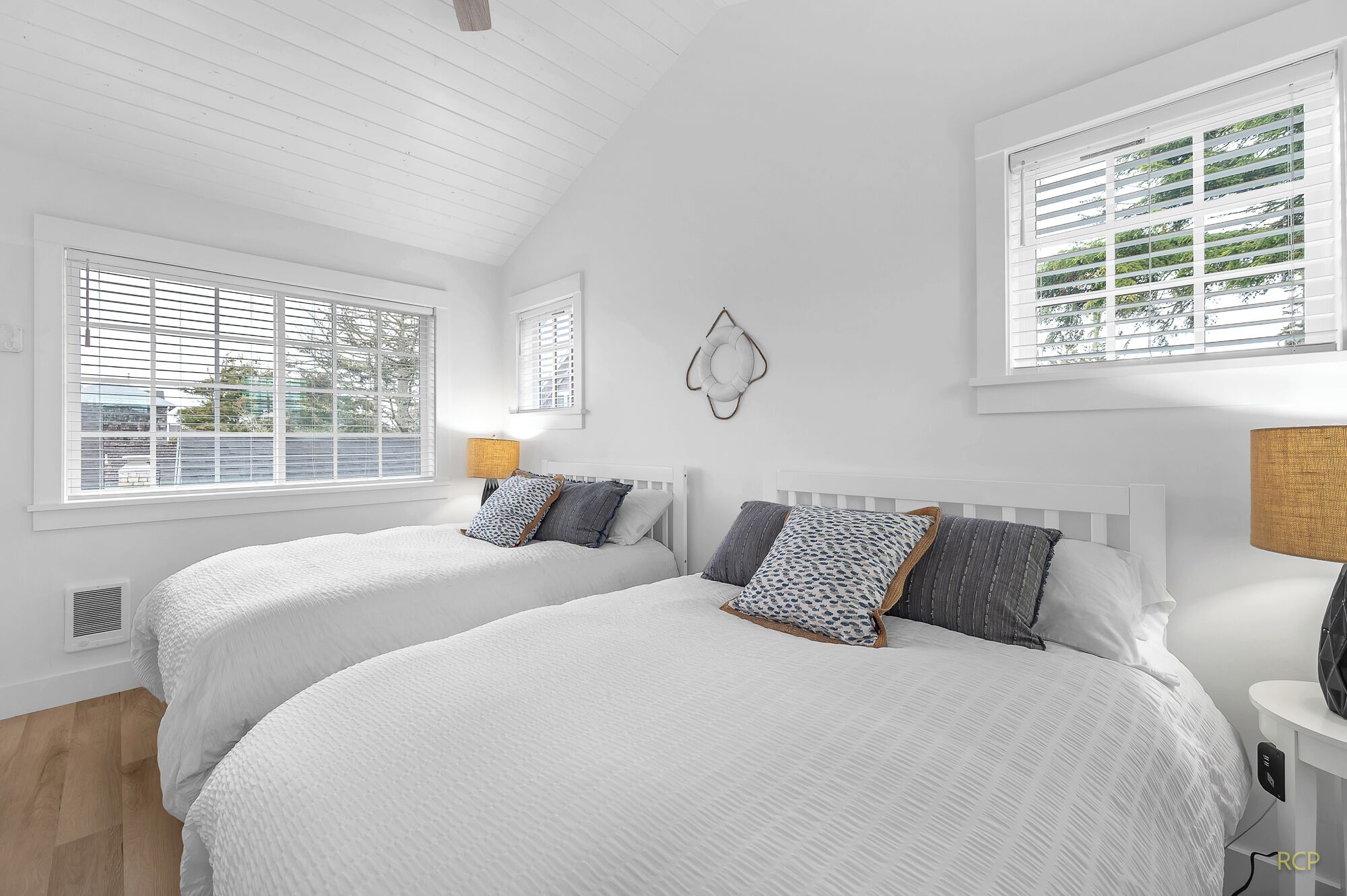 Une chambre avec deux lits, du linge de lit blanc, un plafond en bois et de grandes fenêtres équipées de stores.