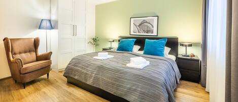 A hotel room with a bed, a chair, a wardrobe, a nightstand, a lamp, and a framed picture on the wall.