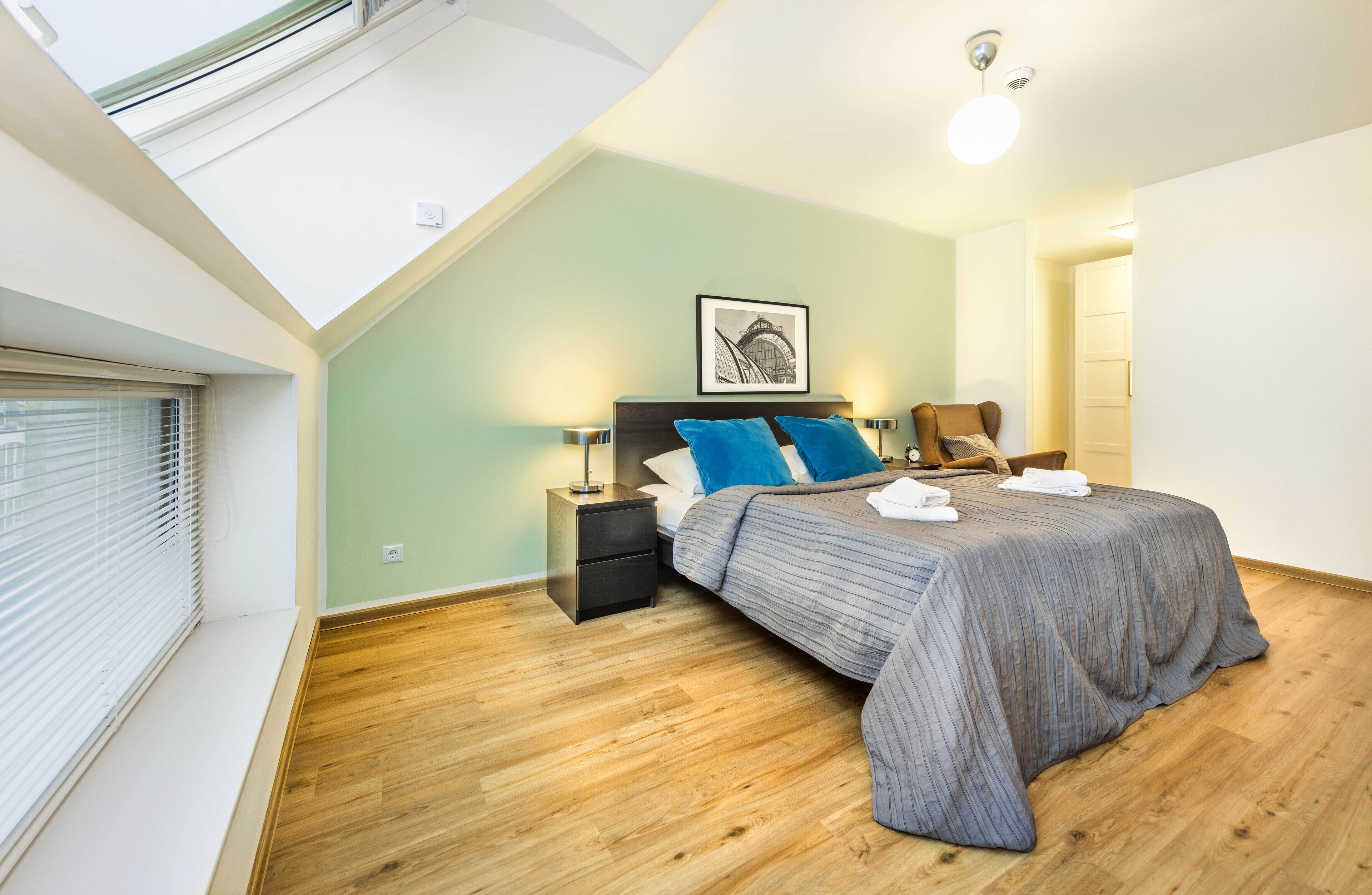 A bedroom with a skylight, a bed with blue bedding, a nightstand, and a chair.