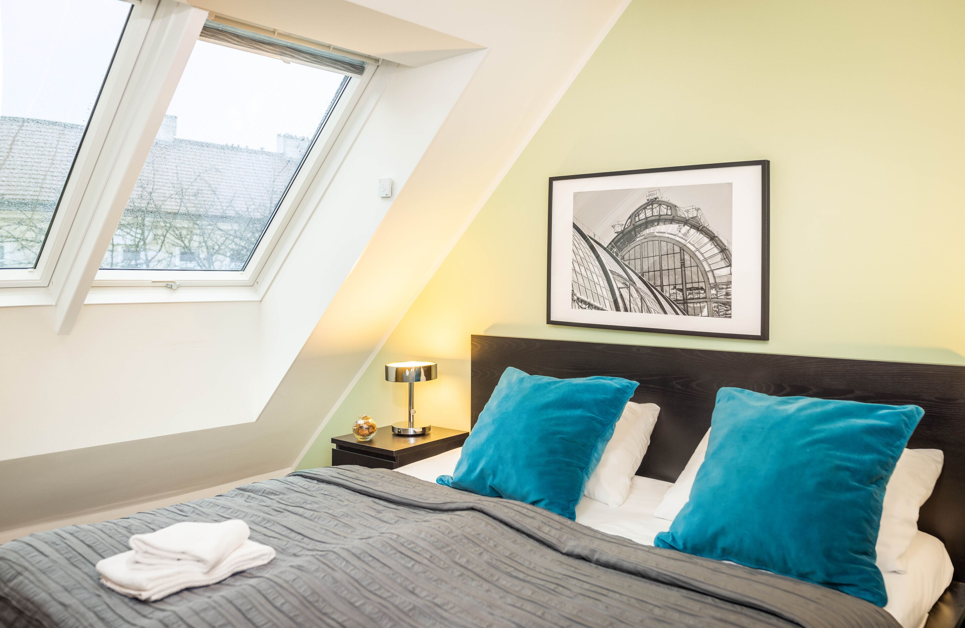 Un dormitorio con cama, almohadas, mesita de noche, lámpara, cuadro enmarcado y ventana.