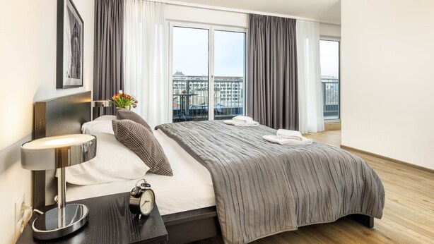 A modern hotel room with a large bed, a bedside table with a lamp, a clock, and a view of the city through large windows. - Jimmy's Apartments Lory (Vienna)