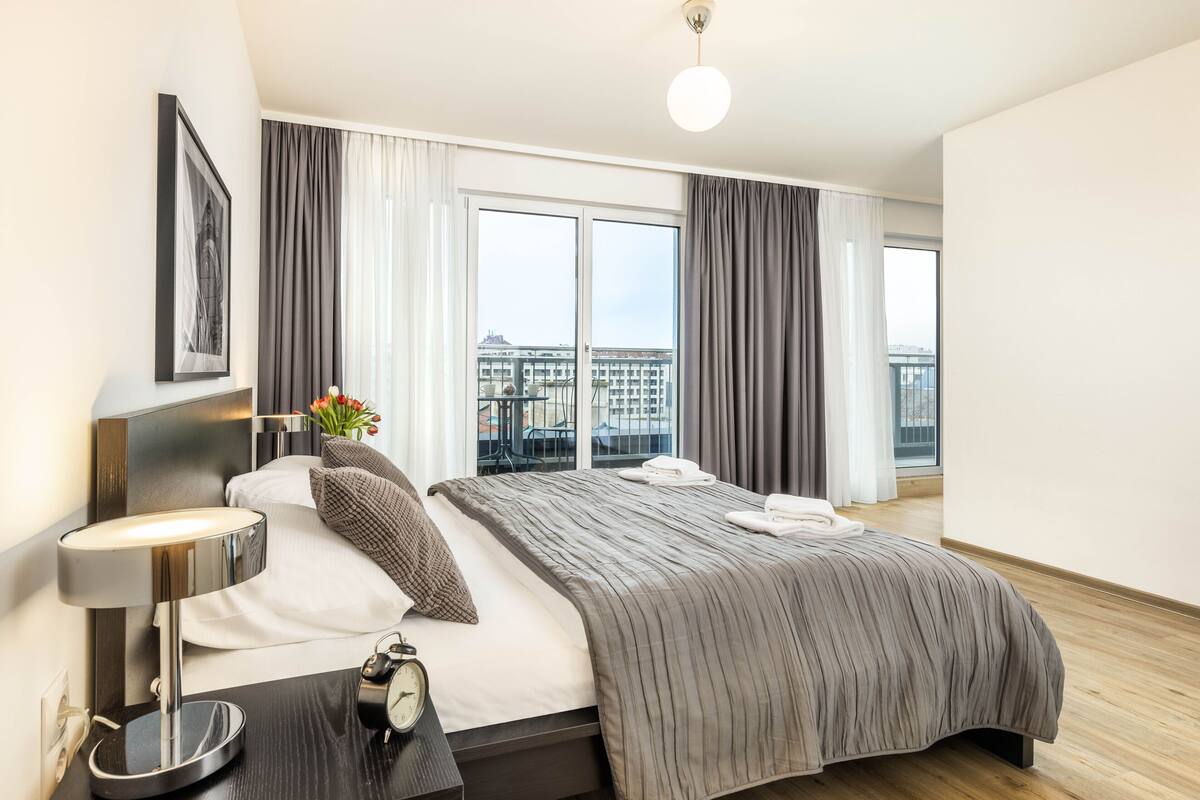 Quarto de hotel moderno com uma cama grande, uma mesa de cabeceira com abajur, relógio e vista da cidade através de amplas janelas.