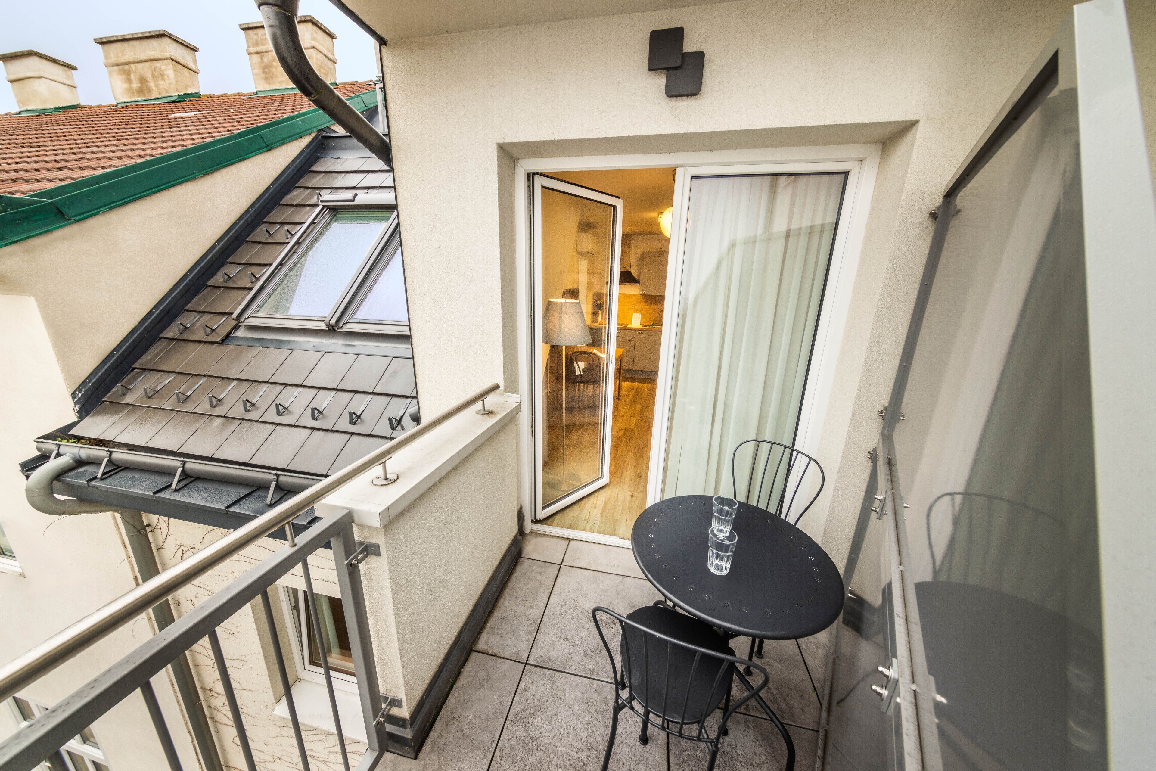 A balcony with a table and chairs, a sliding glass door leading to a kitchen, and a small outdoor area with a tiled floor.