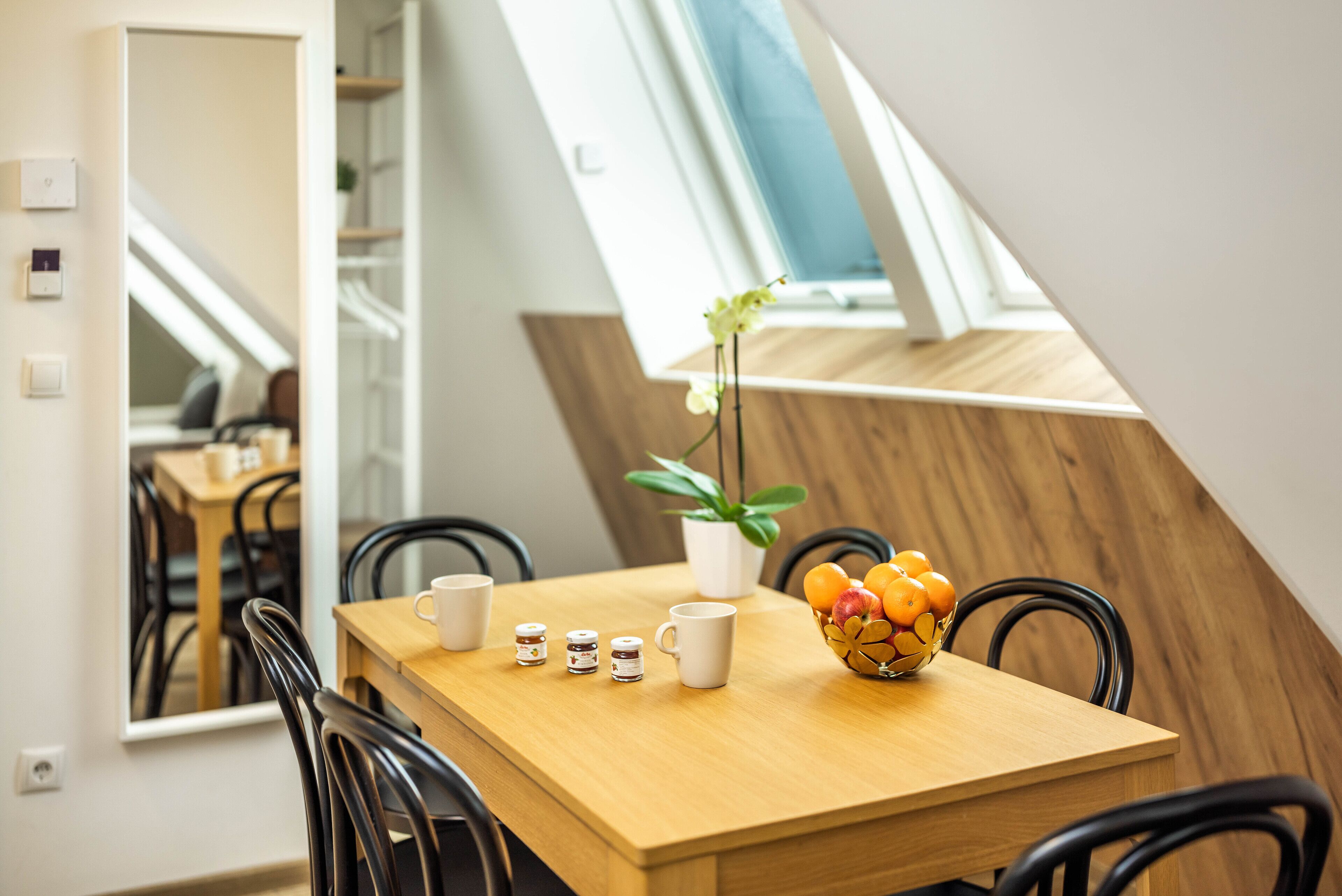 A dining area with a wooden table, black chairs, a white vase with flowers, and a bowl of fruit.