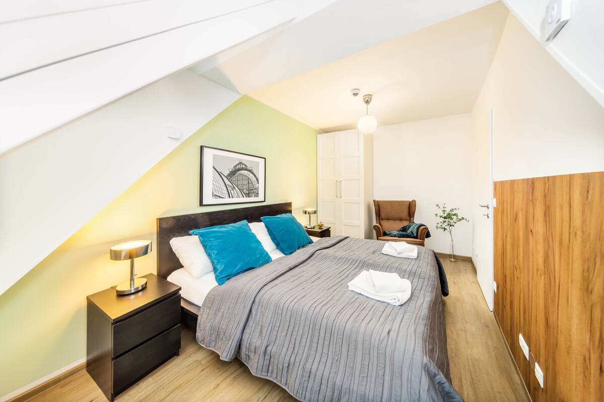 Um quarto moderno com teto inclinado, uma cama com almofadas azuis, uma mesa de cabeceira com abajur, um guarda-roupa de madeira e um quadro na parede.
