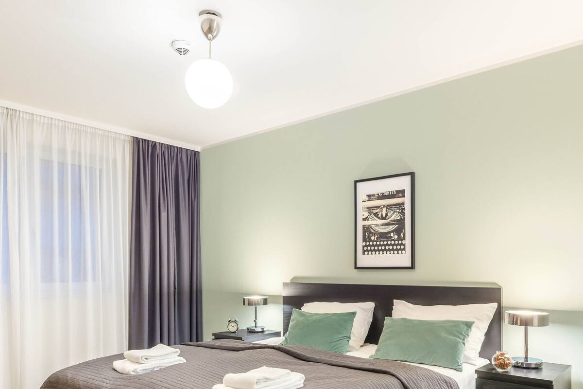 Quarto de hotel moderno com uma cama grande, duas mesas de cabeceira, um criado-mudo, um quadro emoldurado na parede e uma janela com cortinas.