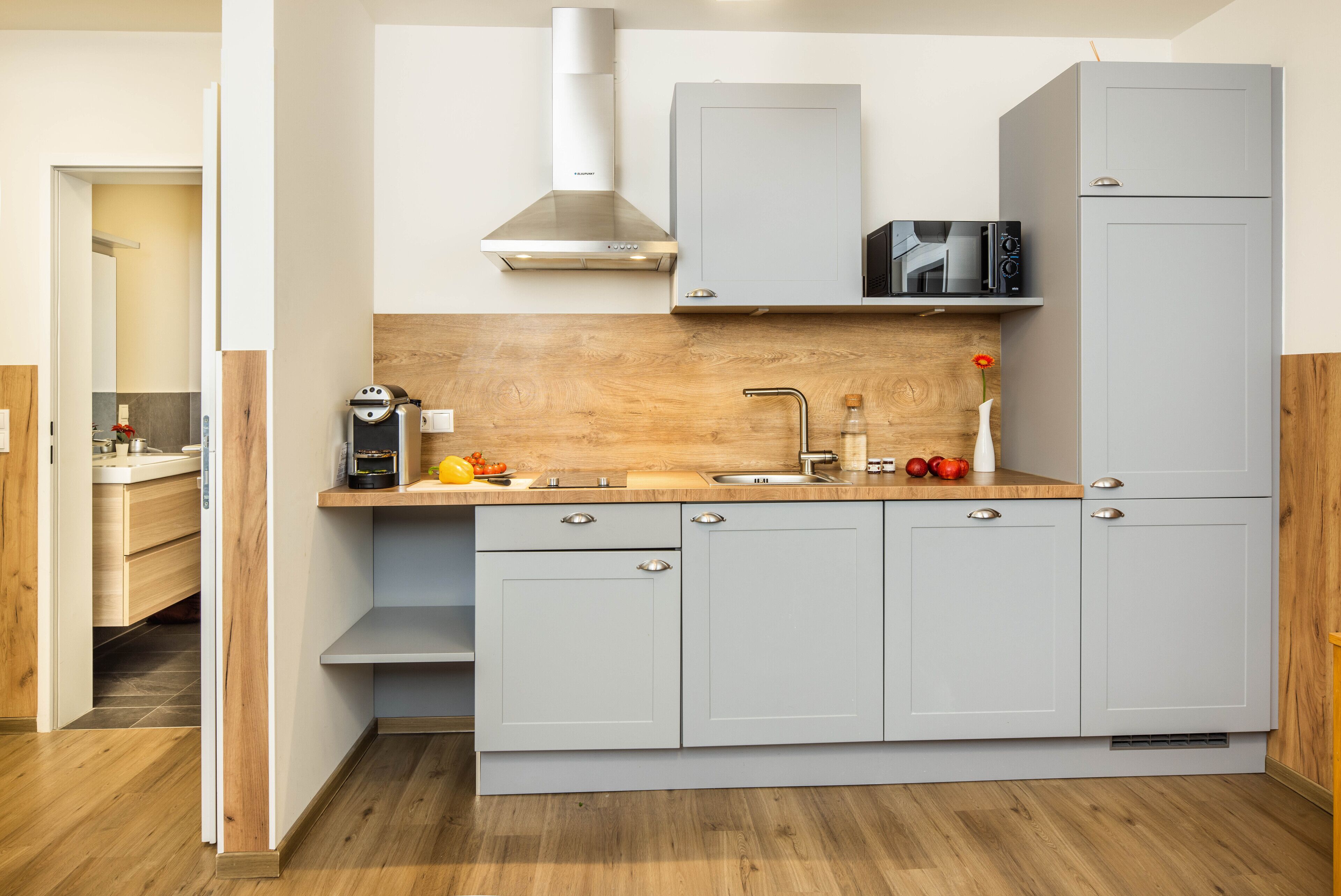 Une cuisine compacte avec des meubles gris, un plan de travail en bois, une hotte en acier inoxydable et un four à micro-ondes.