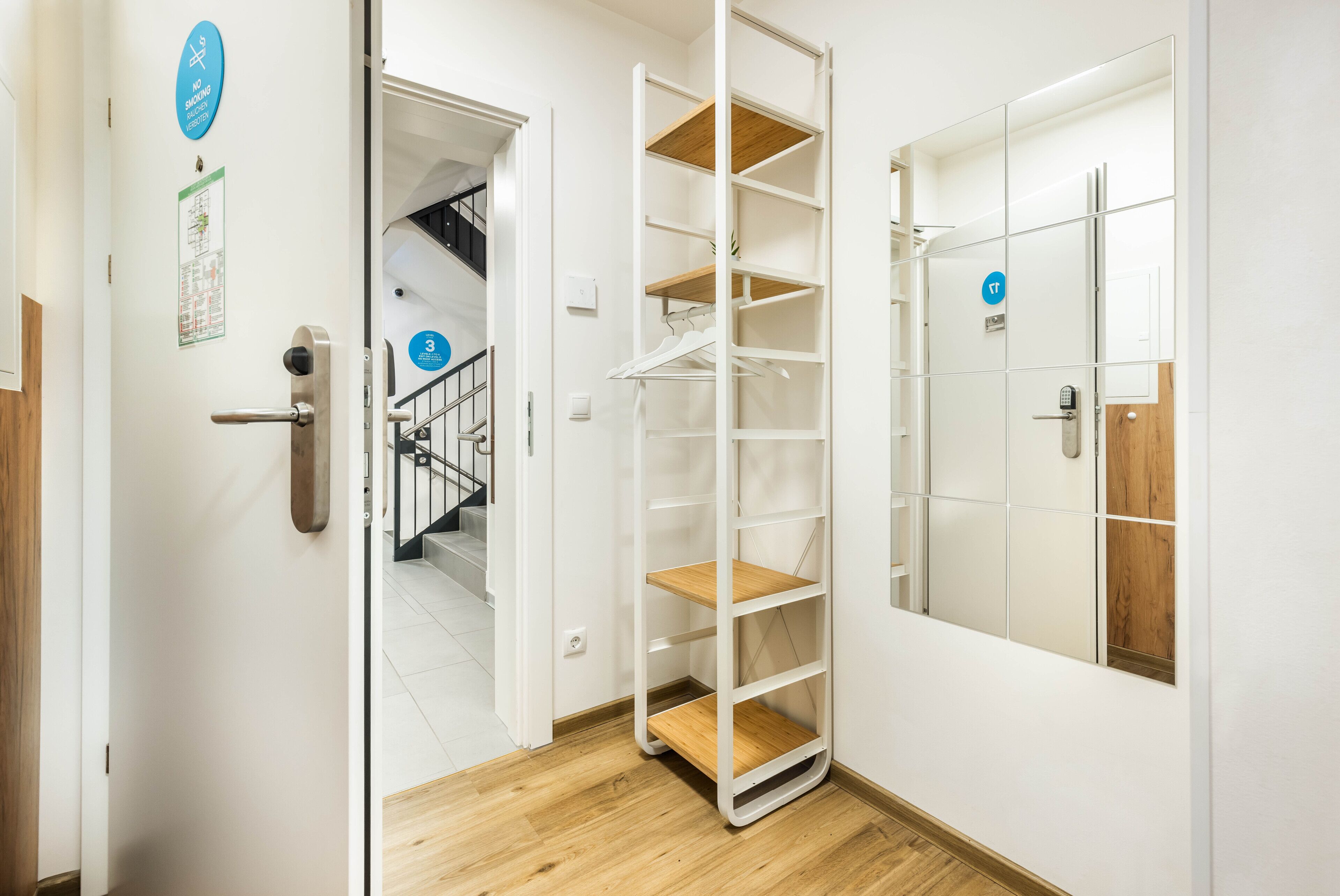 A hotel room with a wooden floor, a white door with a mirror, a metal shelf unit, and a 'No Smoking' sign.