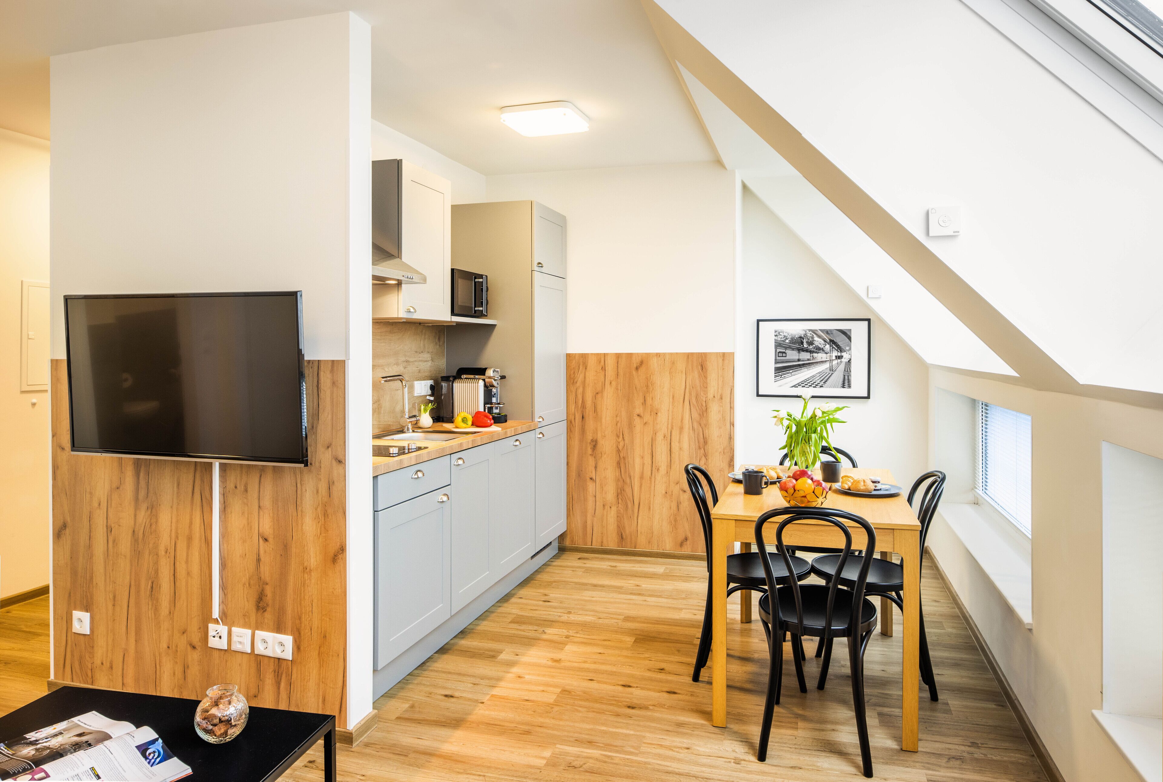 A modern kitchen with a dining area, wooden flooring, and a flat-screen TV mounted on the wall.