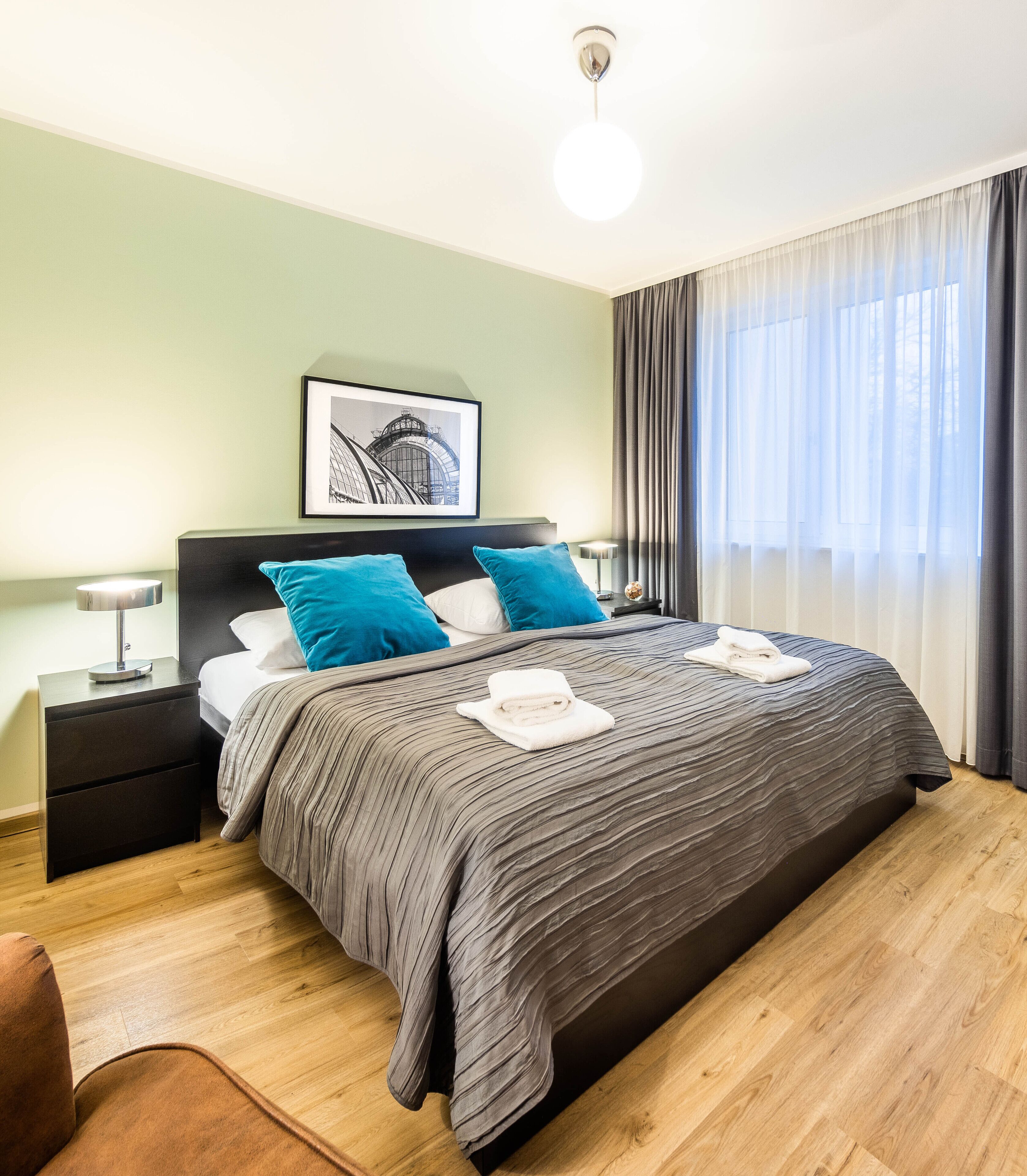 A modern hotel room with a large bed, a nightstand, a lamp, a framed picture on the wall, and a window with curtains.