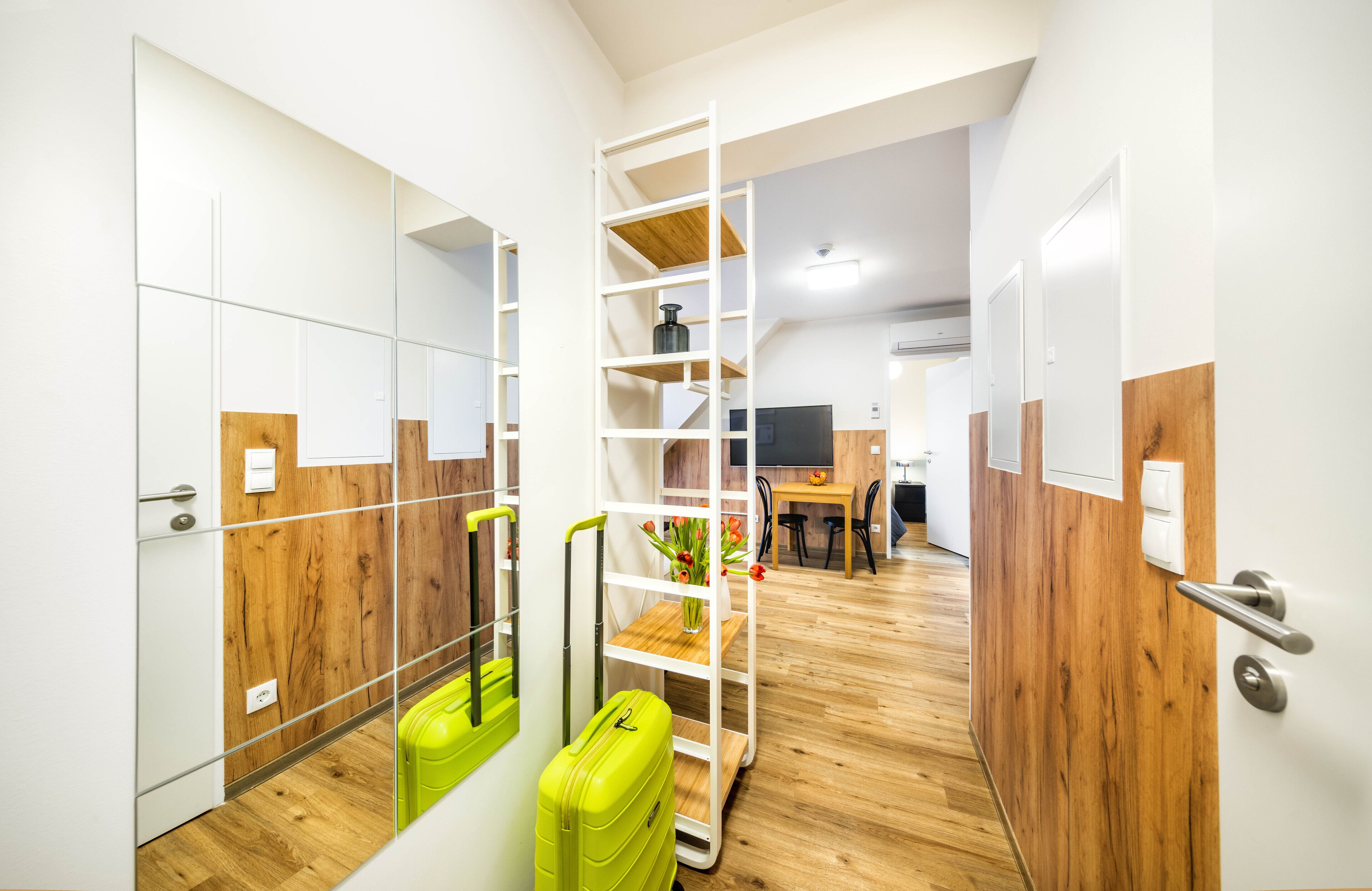 Habitación de hotel moderna con piso de madera, un espejo, una maleta verde y una unidad de estantes de madera.