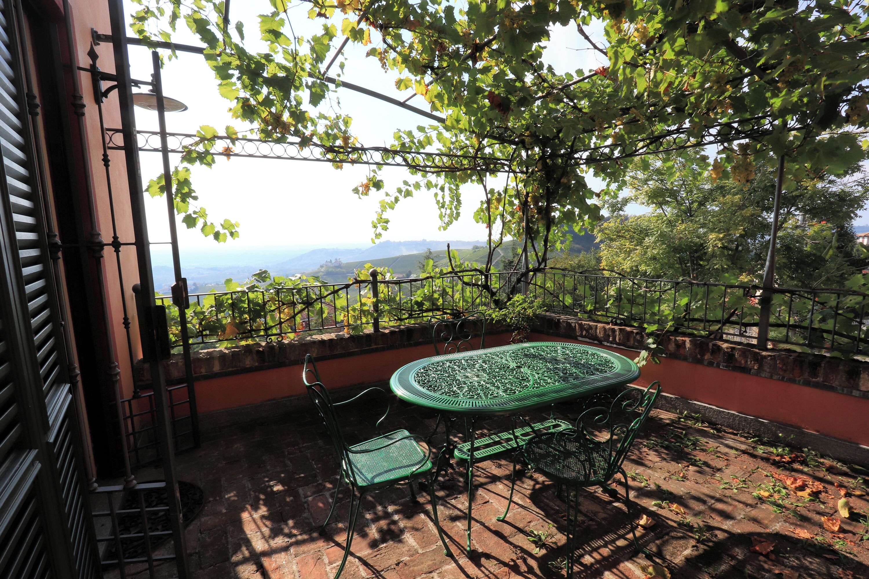 Um pátio com mesa e cadeiras de metal verde, cercado por um muro de tijolos e uma cerca metálica.
