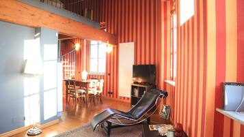 A spacious living area with a black leather chair, a dining table with chairs, a TV, and a bookshelf.