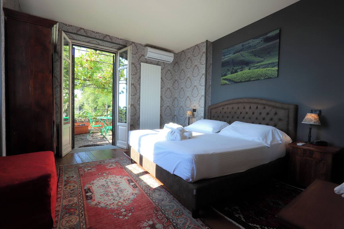 Une chambre avec un grand lit, une table de chevet, un fauteuil rouge et une vue sur un jardin par une porte ouverte.