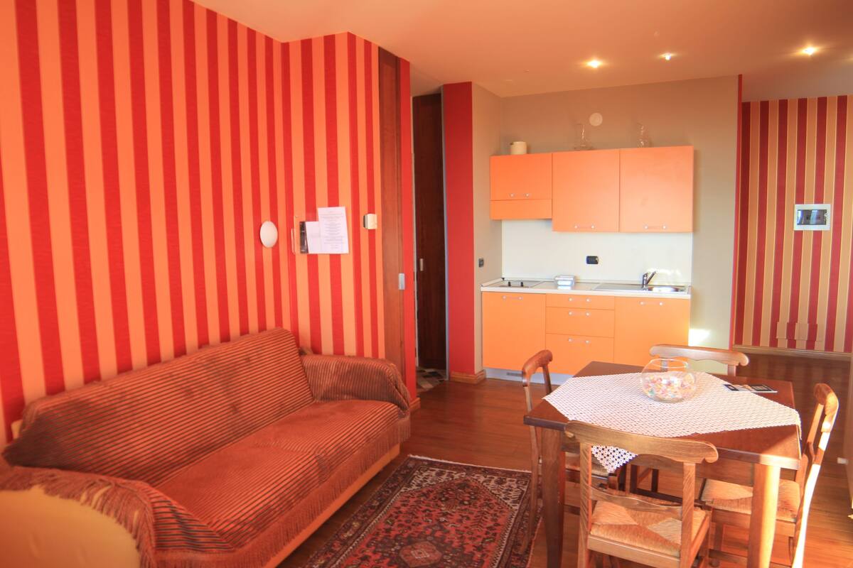 Un salon chaleureux avec un canapé, une table à manger et des chaises, ainsi qu’un coin cuisine doté de meubles orange.