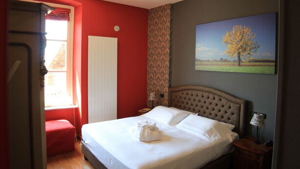 Une chambre d’hôtel avec un lit, un mur rouge, une fenêtre avec des rideaux, un tableau représentant un arbre et un banc rouge.