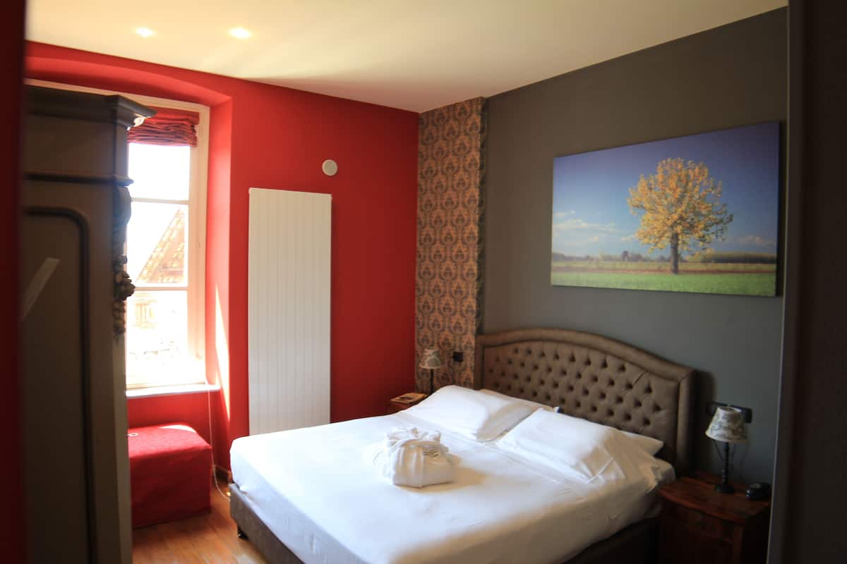 Une chambre d’hôtel avec un lit, un mur rouge, une fenêtre avec des rideaux, un tableau représentant un arbre et un banc rouge.