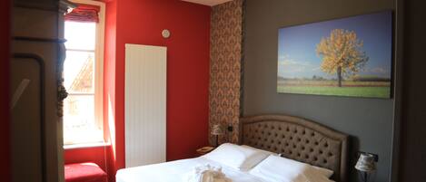 A hotel room with a bed, a red wall, a window with curtains, a painting of a tree, and a red bench.