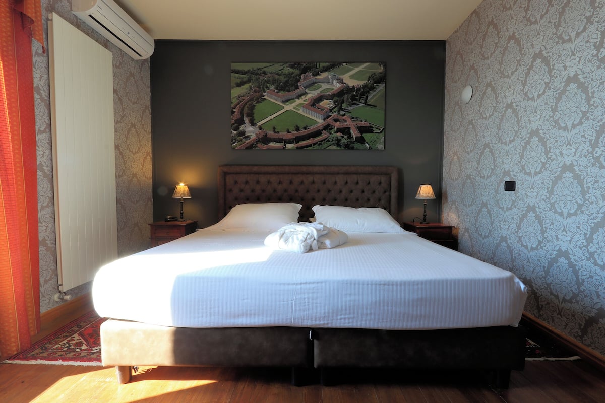 Un lit bien fait, avec une couette blanche et des oreillers, une grande œuvre d’art encadrée au-dessus du lit, et un papier peint à motifs sur les murs.