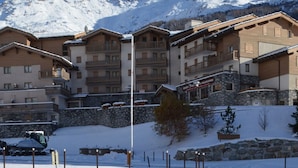 A snow-covered mountain resort with multi-story buildings, balconies, and a clear view of the mountains in the background.