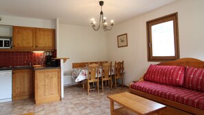 A compact kitchen with wooden cabinets, a dining table with chairs, a sofa, and a window with a view.