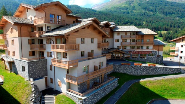A multi-story hotel with balconies, a stone facade, and a view of mountains.