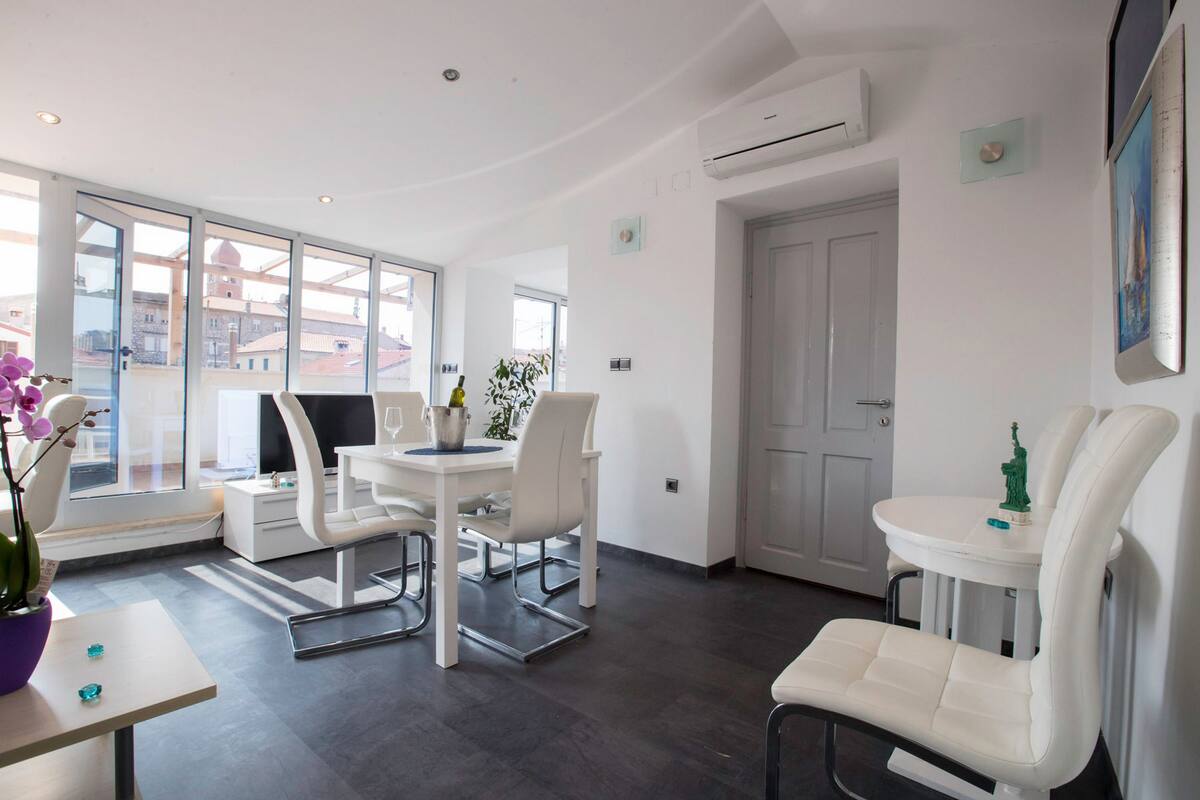 Une pièce moderne et bien éclairée, comprenant un coin repas, un espace salon et une grande fenêtre donnant sur des immeubles.