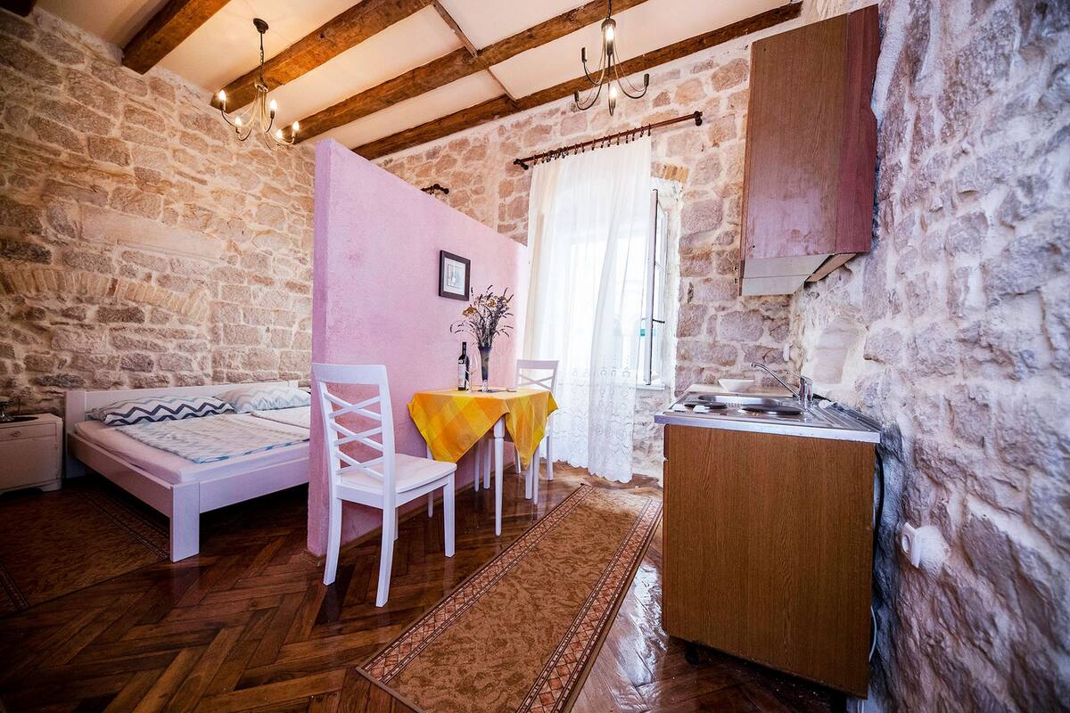 Une pièce rustique comprenant un lit, une table à manger avec des chaises, un petit coin cuisine et des murs en pierre.