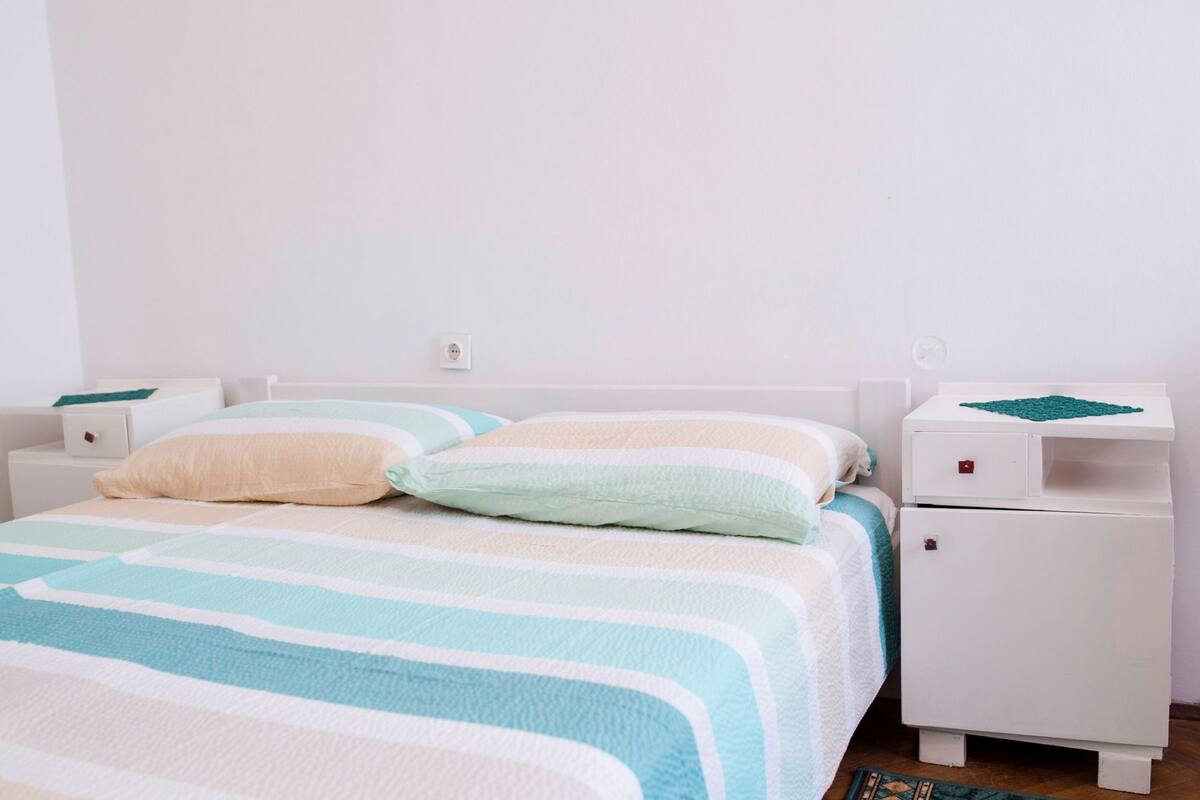 Un lit bien fait, avec une literie à rayures, une table de chevet blanche dotée d’un tiroir, et une petite table de chevet blanche sur laquelle est posée une nappe verte.