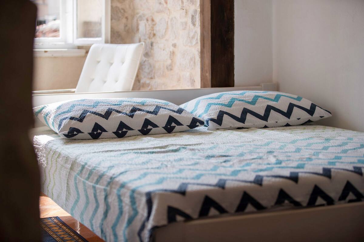 Un lit avec des coussins à motifs et une couverture rayée, une chaise blanche et, en arrière-plan, un mur en pierre.