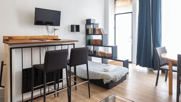 A modern living space with a flat-screen TV mounted on the wall, a wooden counter, two black chairs, a small bed with a blanket, a bookshelf, and a dining table with chairs. - Souplex - 4P - Paris city center (Paris)