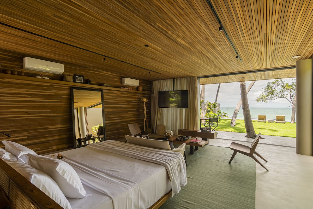 Une chambre moderne avec un grand lit, un bureau, une chaise et un téléviseur. On peut admirer l’océan et les palmiers depuis la fenêtre.