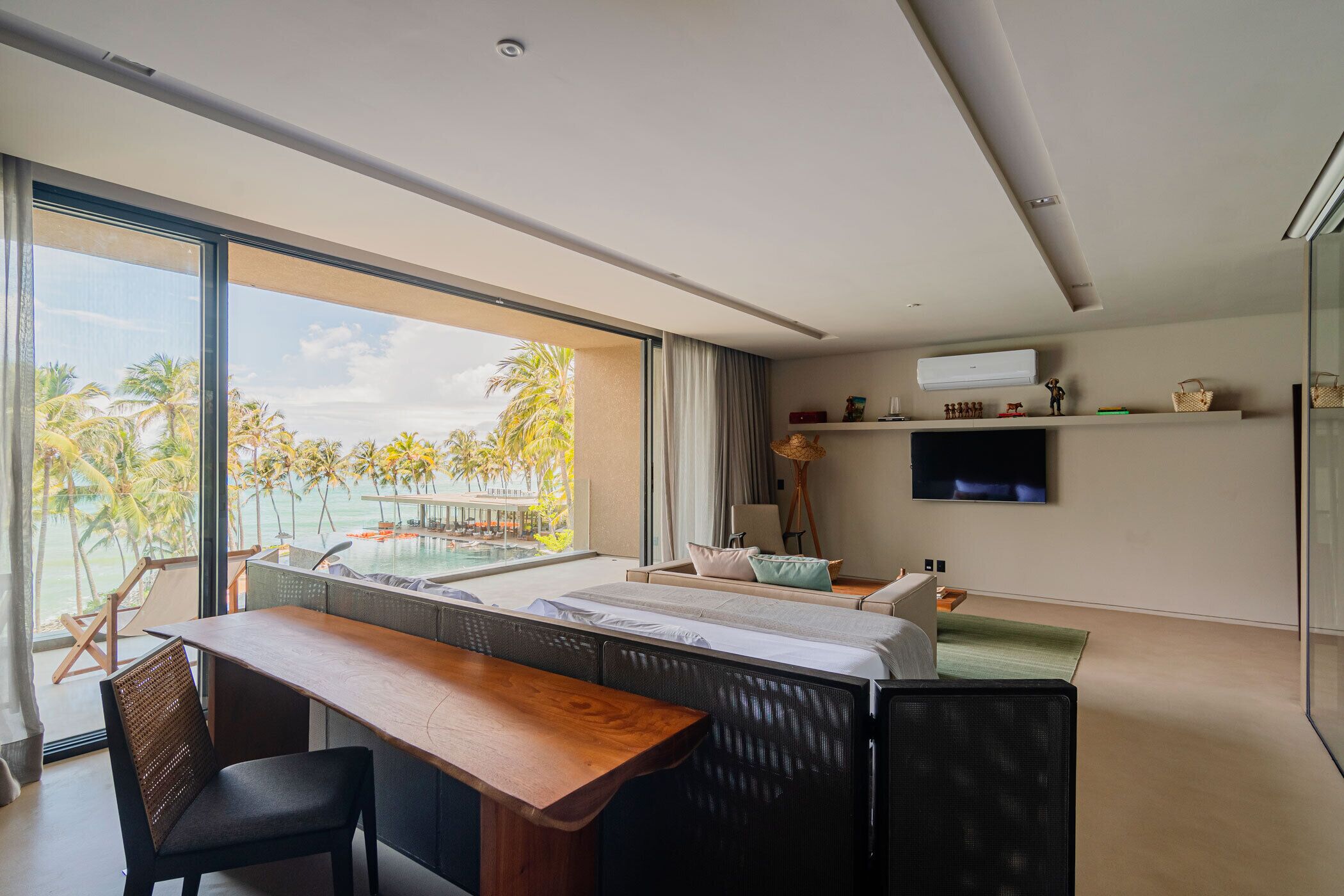 Une chambre d’hôtel moderne dotée d’une grande fenêtre donnant sur une piscine et des palmiers, un lit avec vue, un bureau avec une chaise et un téléviseur à écran plat fixé au mur.