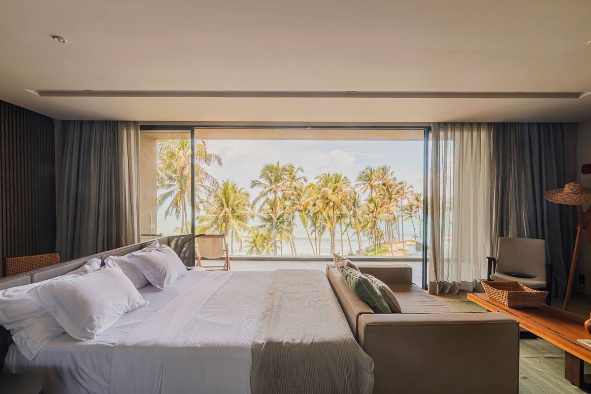 Une chambre d’hôtel avec un grand lit, un canapé, un bureau et une vue sur les palmiers et la mer.