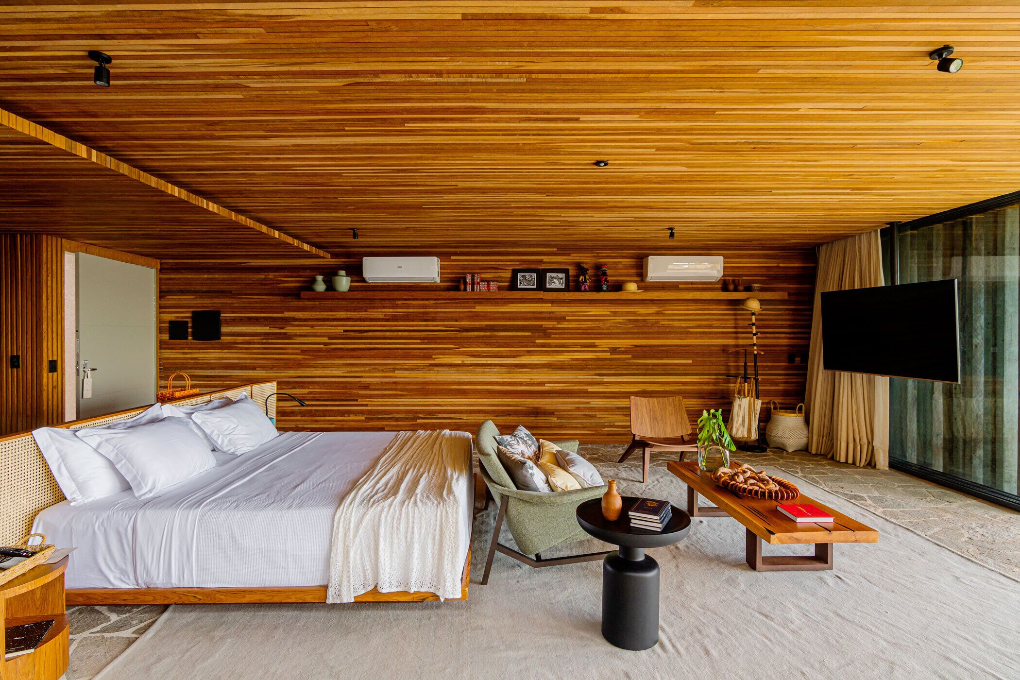 A modern bedroom with a wooden ceiling, a large bed, a green armchair, a small table with a vase, and a TV on the right.