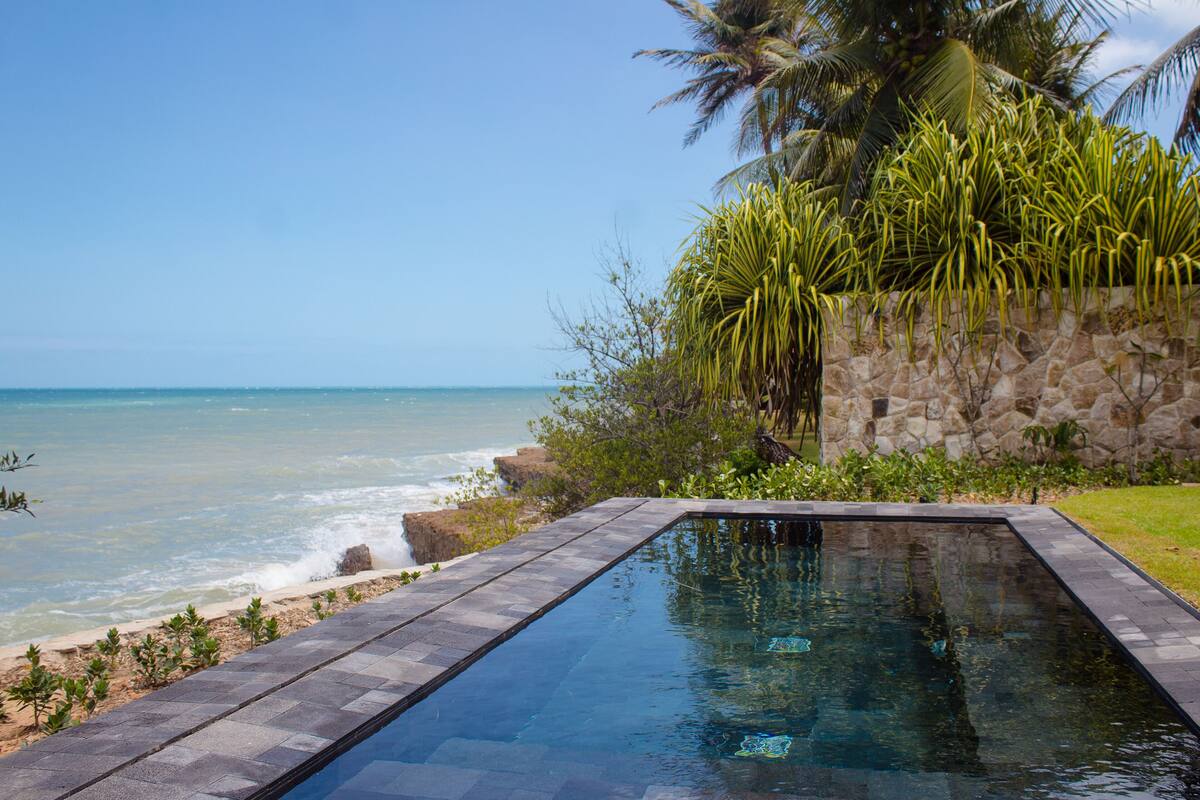 Une piscine avec vue sur l’océan et un mur de pierre orné de plantes tropicales.