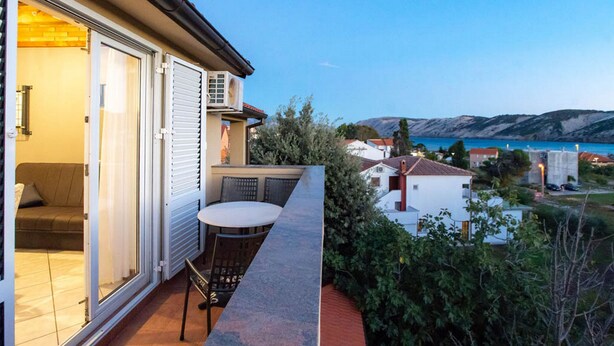A balcony with a table and chairs, overlooking a coastal area with houses and hills. - Apartment Sanic Rab (Lopar)