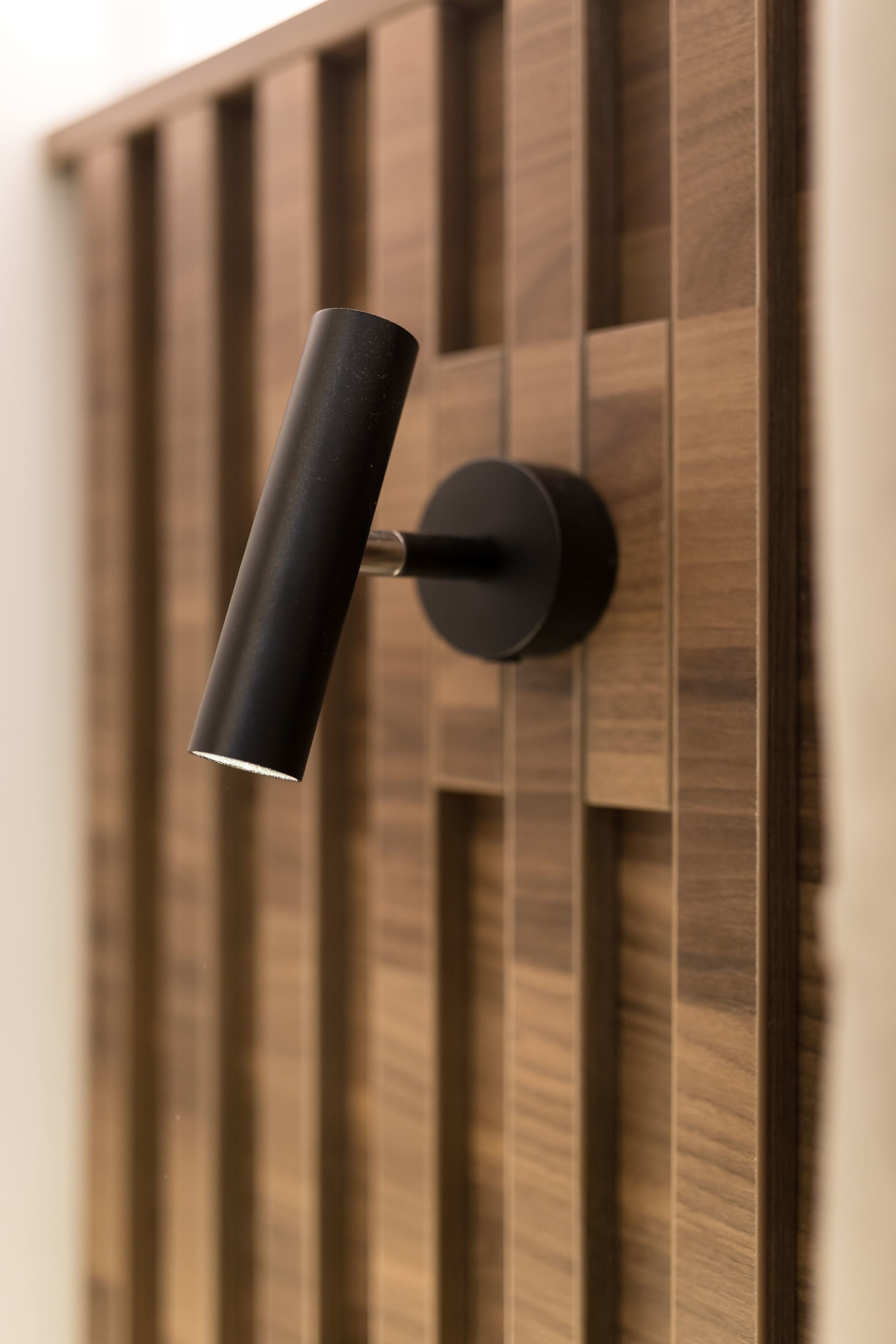 A modern wall-mounted light fixture with a cylindrical shade and a round base, set against a wooden panel background.