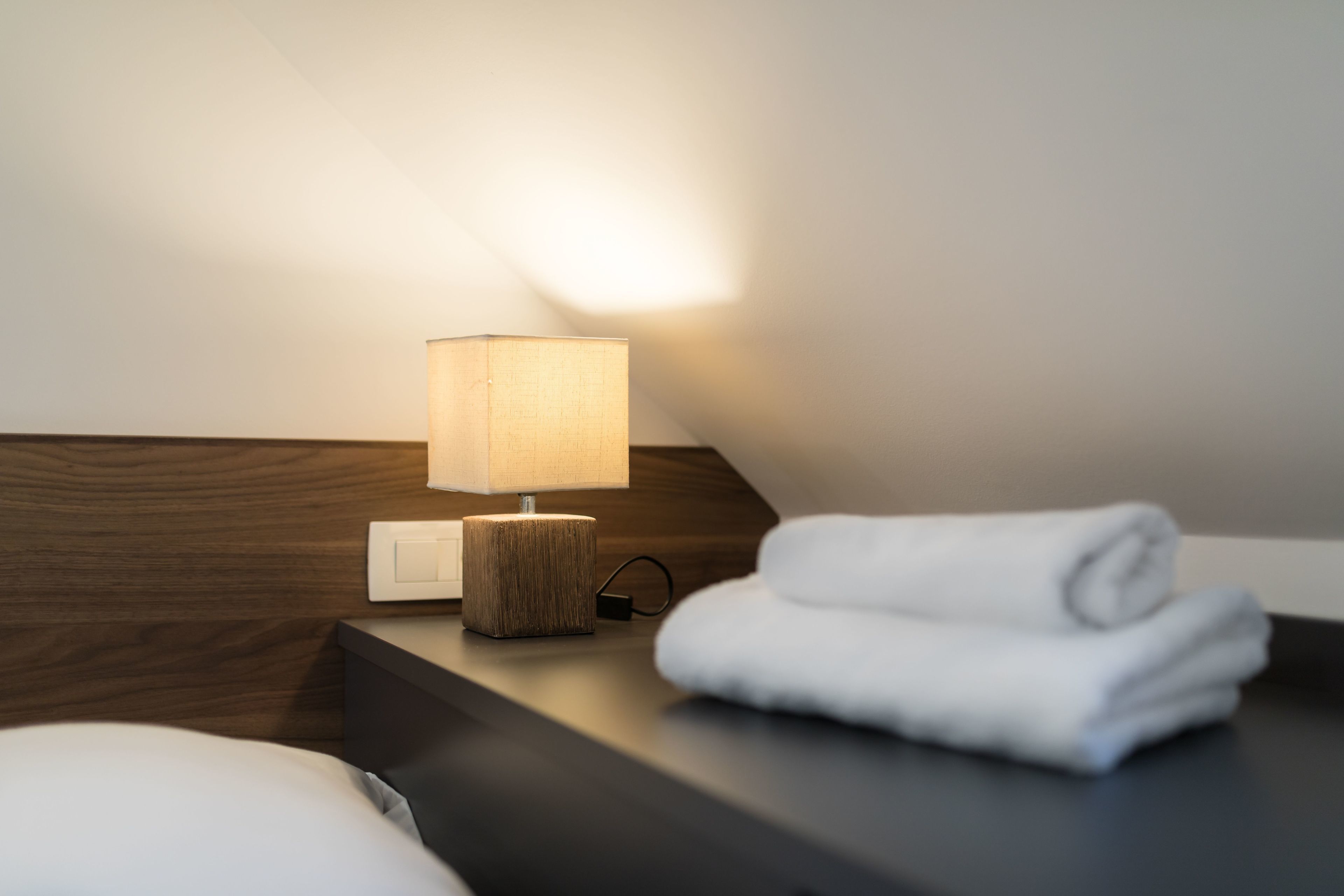 A neatly made bed with a white pillow and a folded white towel on a dark wooden nightstand.